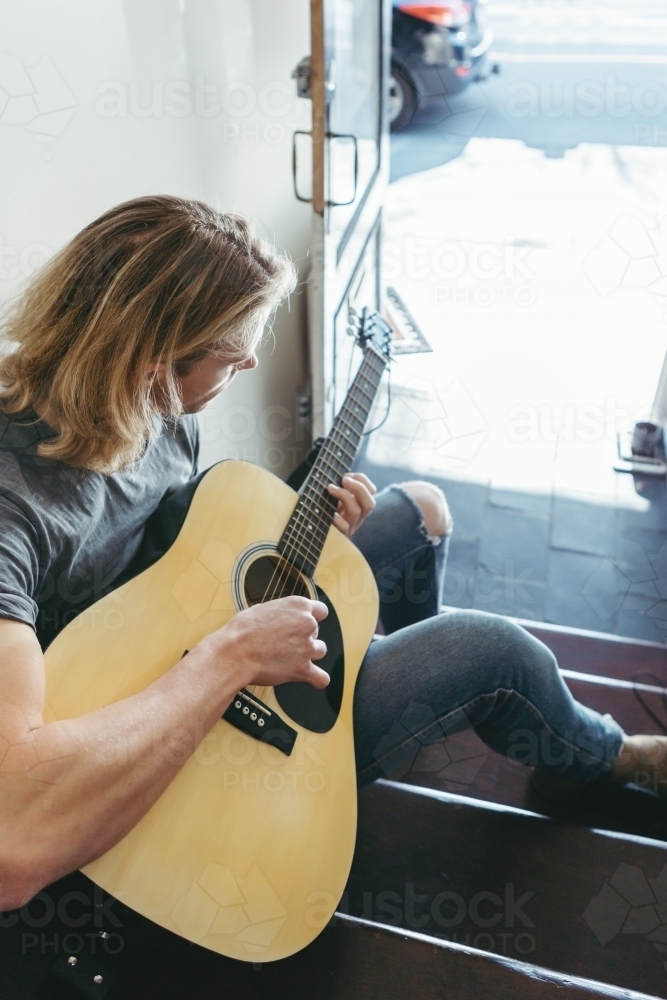 Blonde musician playing guitar in apartment stairs - Australian Stock Image