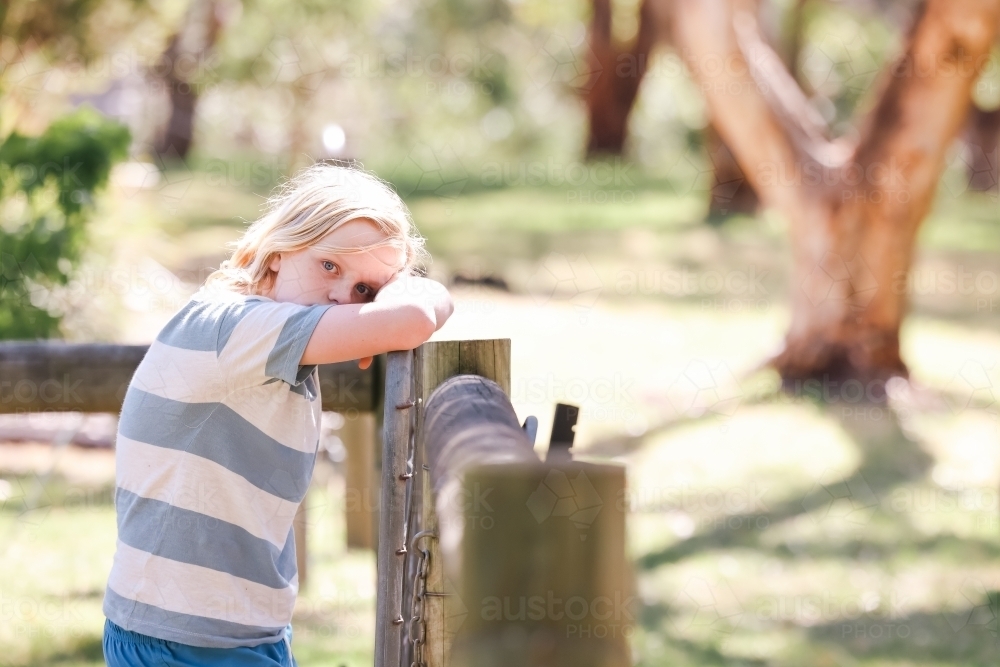 Blonde pre-teen boy leaning on fence with serious expression - Australian Stock Image