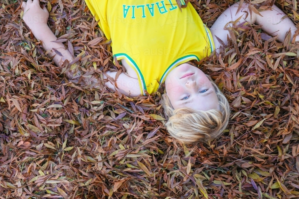 Blonde pre-teen boy lying on in fallen leaves with serious expression - Australian Stock Image
