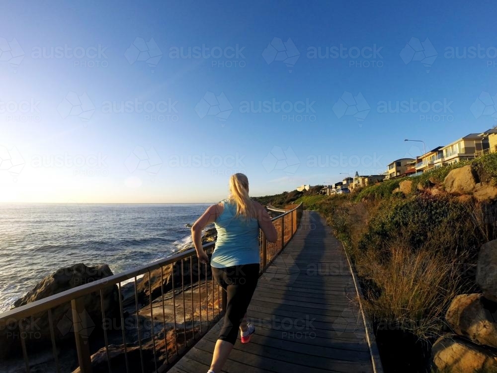 Blonde woman running along ocean boardwalk at sunrise - Australian Stock Image