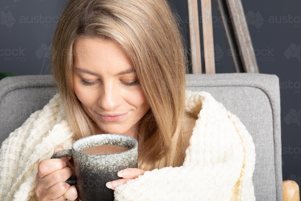 Blonde young lady wrapped in cream blanket hugging a mug - Australian Stock Image