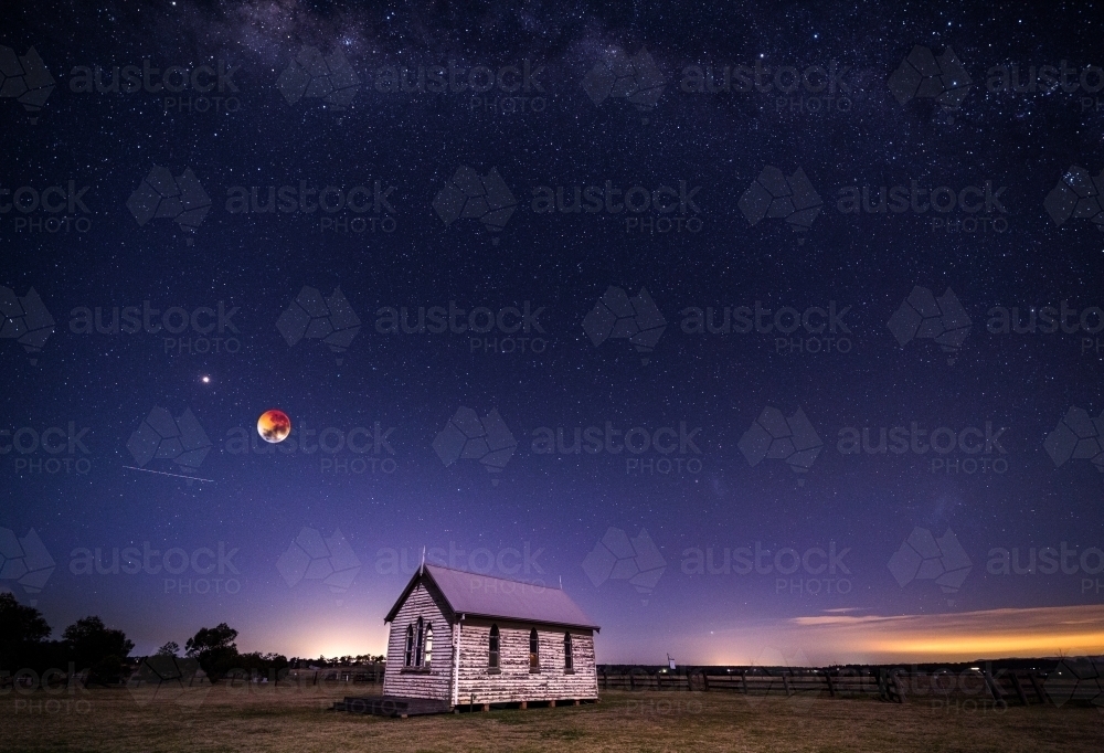 Blood moon setting, planet mars and a universe of stars shining brightly above the rustic chapel - Australian Stock Image