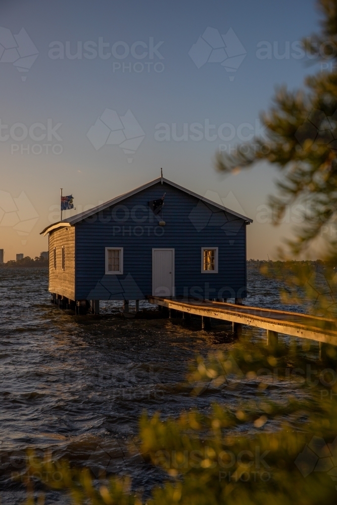 Blue boat house with walkway under clear skies with blurry leaves in the front - Australian Stock Image