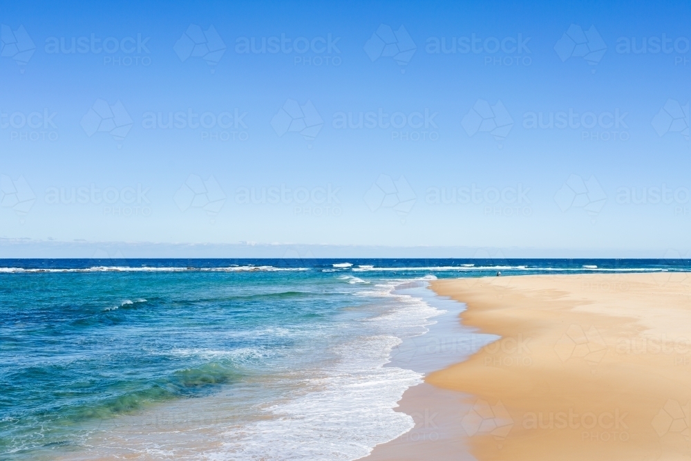 Blue sky above turquoise ocean water and white waves washing up to golden sand - Australian Stock Image