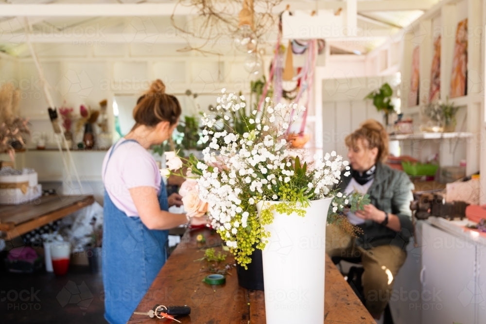 Blurred female florist and assistant at work bench with vase of baby's breath flowers in foreground - Australian Stock Image