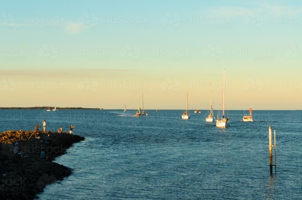 Boats and yachts returning to harbour with people fishing on rocks - Australian Stock Image