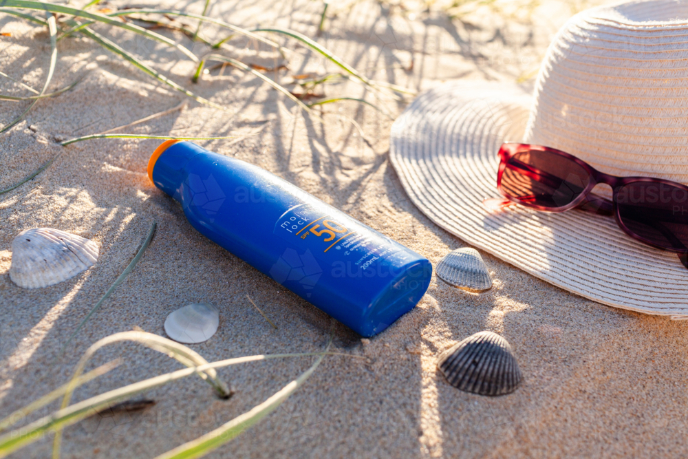 Bottle of 50+ SPF sunscreen on beach sand by sun hat and sunglasses - Australian Stock Image