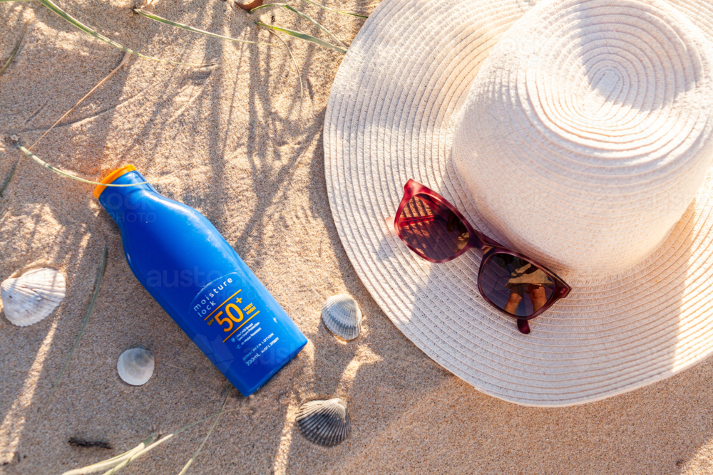 Bottle of 50+ SPF sunscreen on beach sand by sun hat and sunglasses - Australian Stock Image