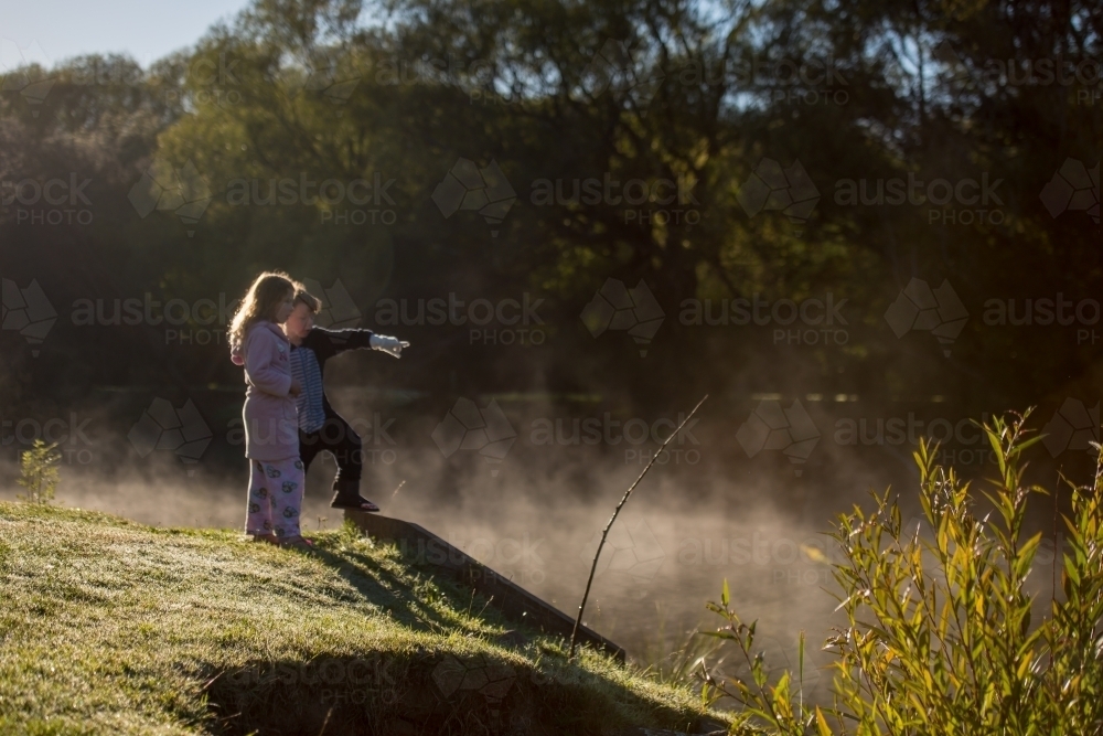 Boy and girl rugged up on cold morning fog day watching ducks in river - Australian Stock Image