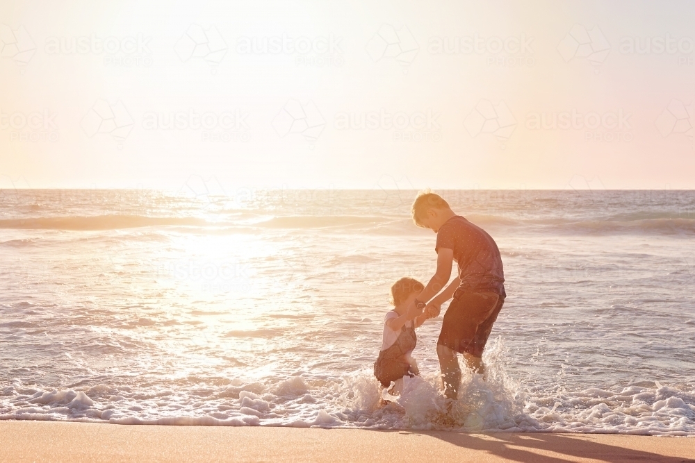 Boy and Toddler Jumping in the Waves on a Beach at Sunset - Australian Stock Image