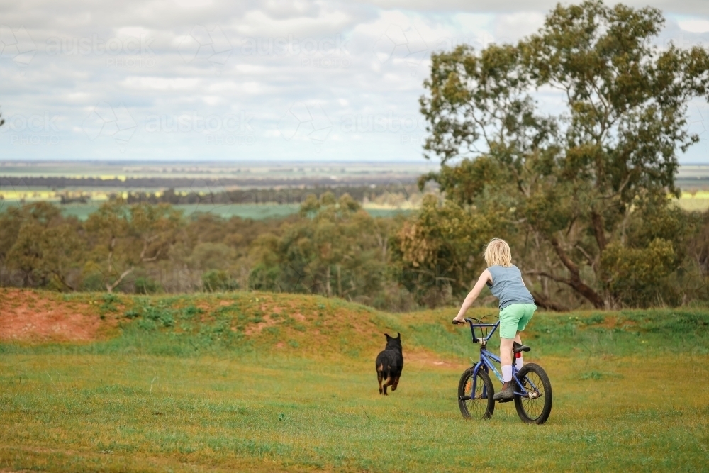 Boy bike riding with rottweiler dog on grass with view of farmland in the distance - Australian Stock Image