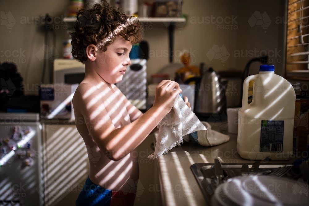 Boy doing dishes in kitchen with light from vertical blinds - Australian Stock Image