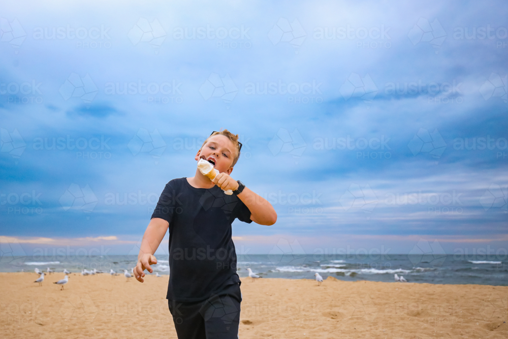 Boy enjoying ice cream on beach with seagulls and blue sky in background - Australian Stock Image