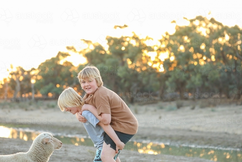 Boy giving brother a piggy back near creek with pet lamb watching - Australian Stock Image