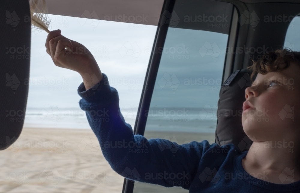 Boy holding a feather out the window of a car next to the beach. - Australian Stock Image