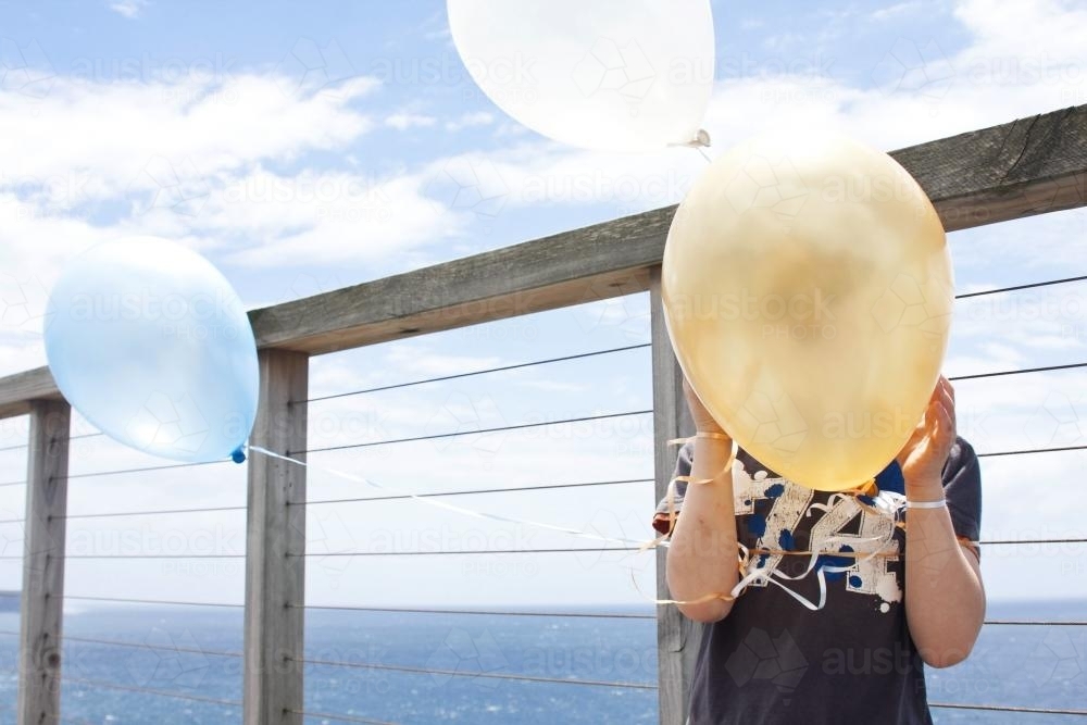 Boy holding balloons in his face by the sea - Australian Stock Image
