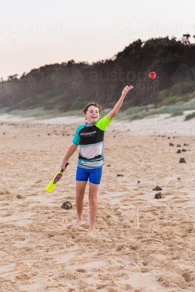 Image of Boy holding cricket bat throwing ball happy having fun on sand at  beach - Austockphoto