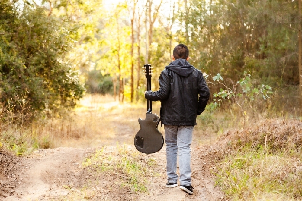 Boy holding guitar walking away through bushland - Australian Stock Image