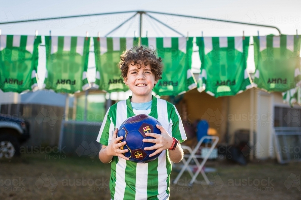 Boy holding soccer ball with team shirts hanging behind - Australian Stock Image