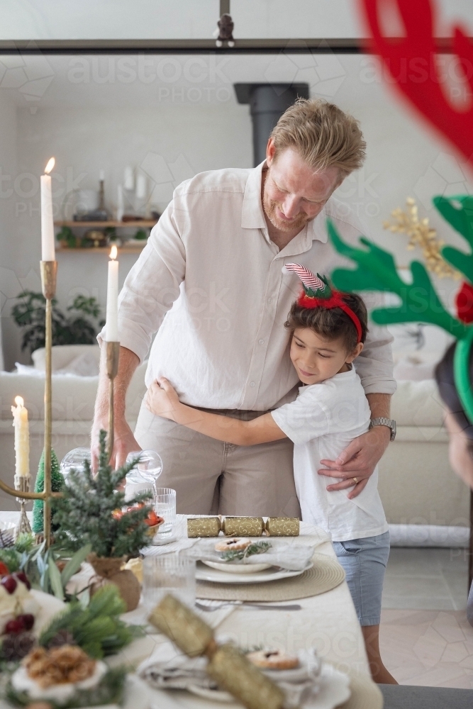 Boy hugging dad pouring water at Christmas table - Australian Stock Image