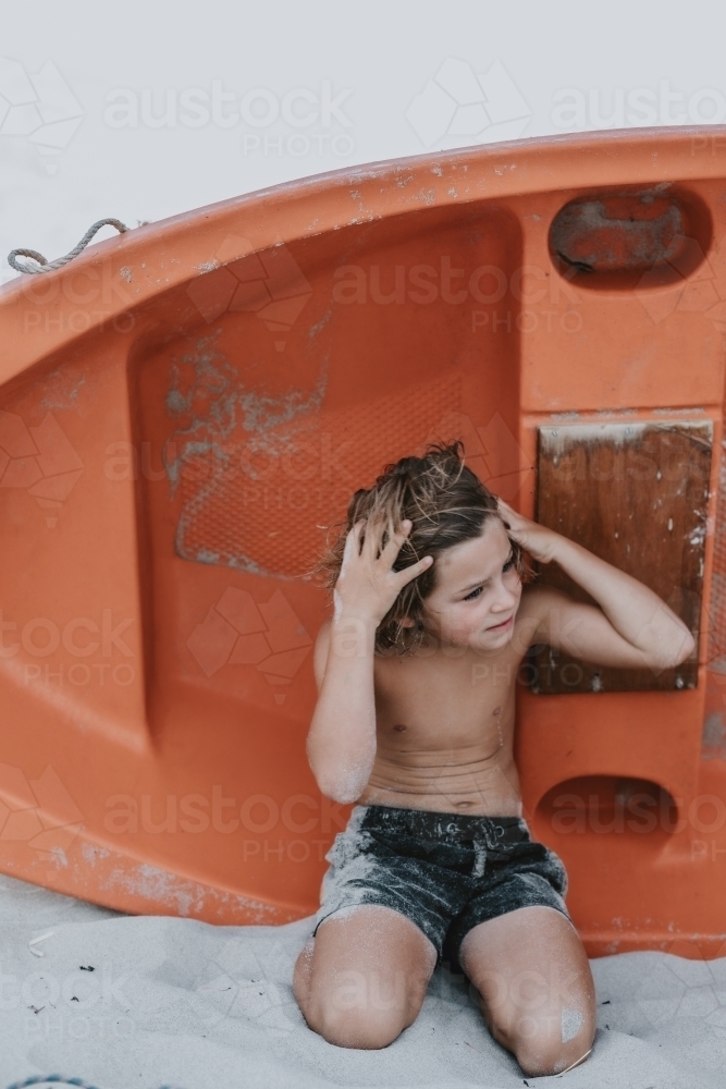 Boy in his swimmers kneeling in the sand in front of a red row boat - Australian Stock Image