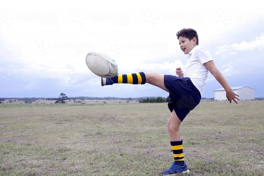 Boy kicking a rugby football in a field - Australian Stock Image