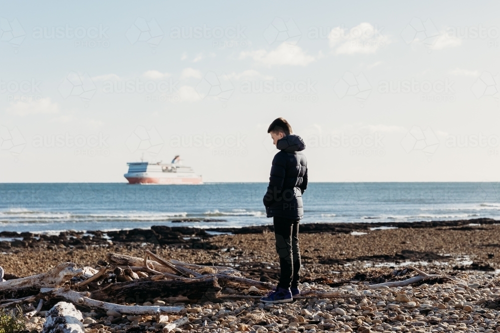 Boy looking at driftwood on beach with cruise ship in background - Australian Stock Image