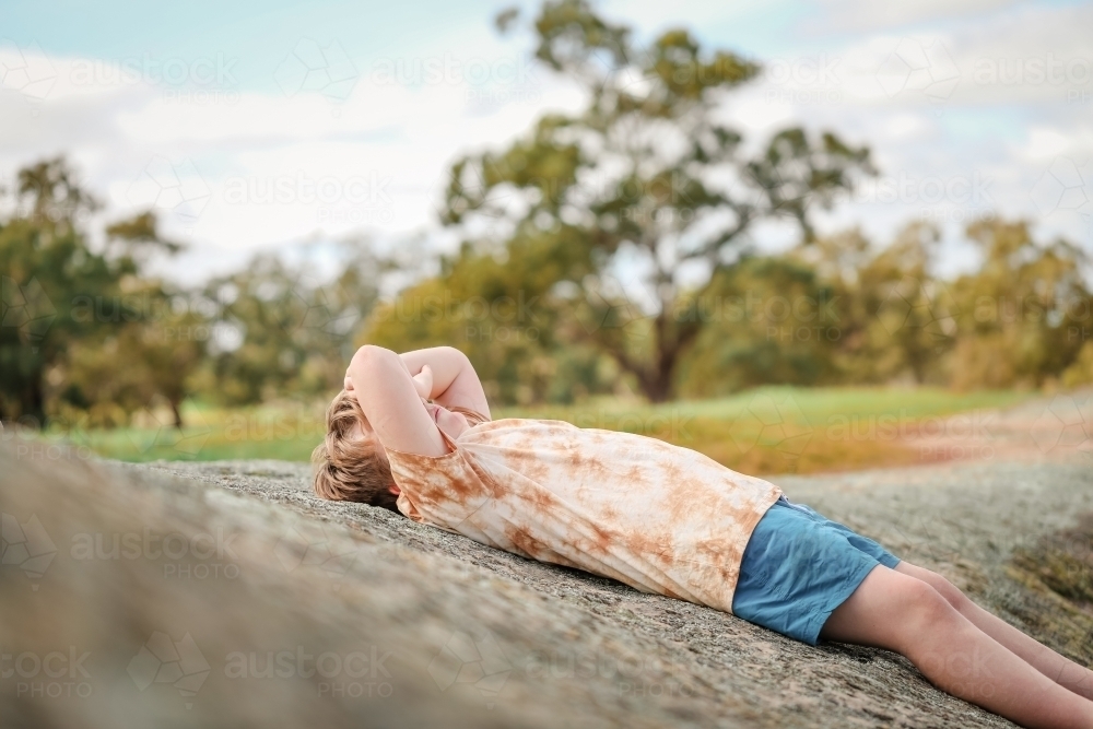 Image of Boy lying outdoors on rock covering face from sun - Austockphoto