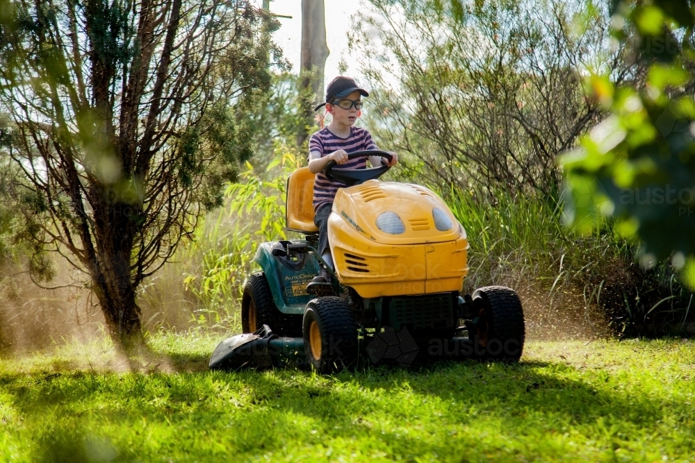 Boy mowing the front yard on a ride on lawn mower - Australian Stock Image