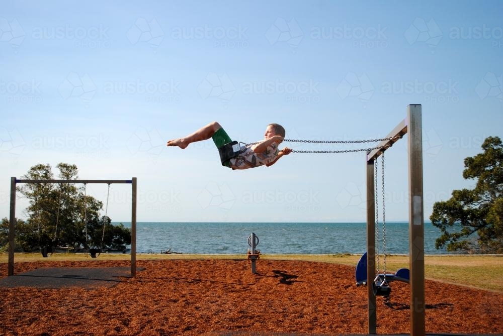 Boy on a swing in a playground near the sea - Australian Stock Image