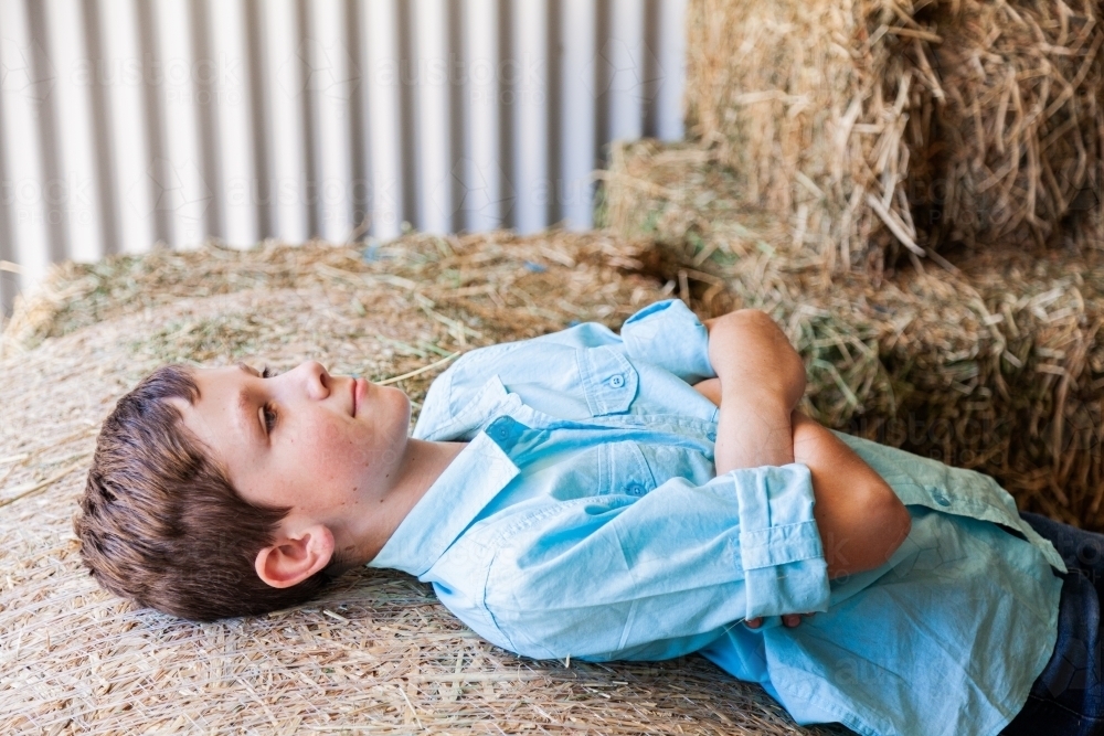 Boy on farm relaxing on hay bale in shed - Australian Stock Image