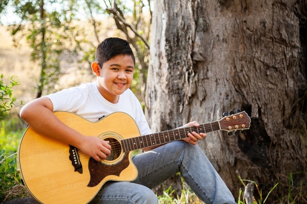 Boy playing country music on acoustic guitar in bushland - Australian Stock Image