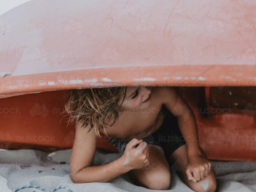 Boy playing hide and seek under red boat on beach - Australian Stock Image