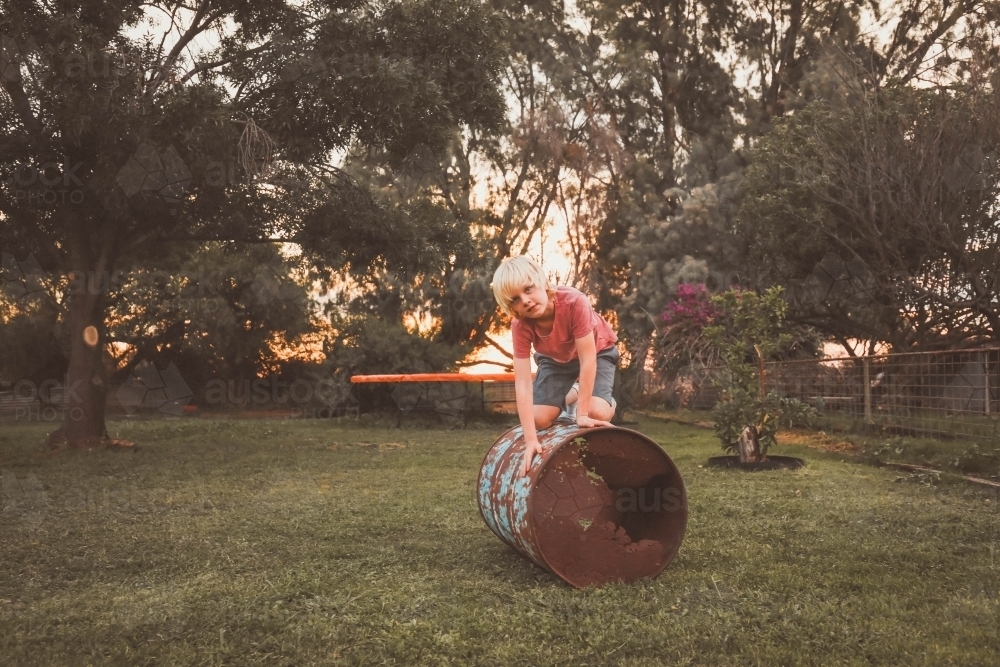 Boy playing on rusty metal drum in country backyard on farm - Australian Stock Image