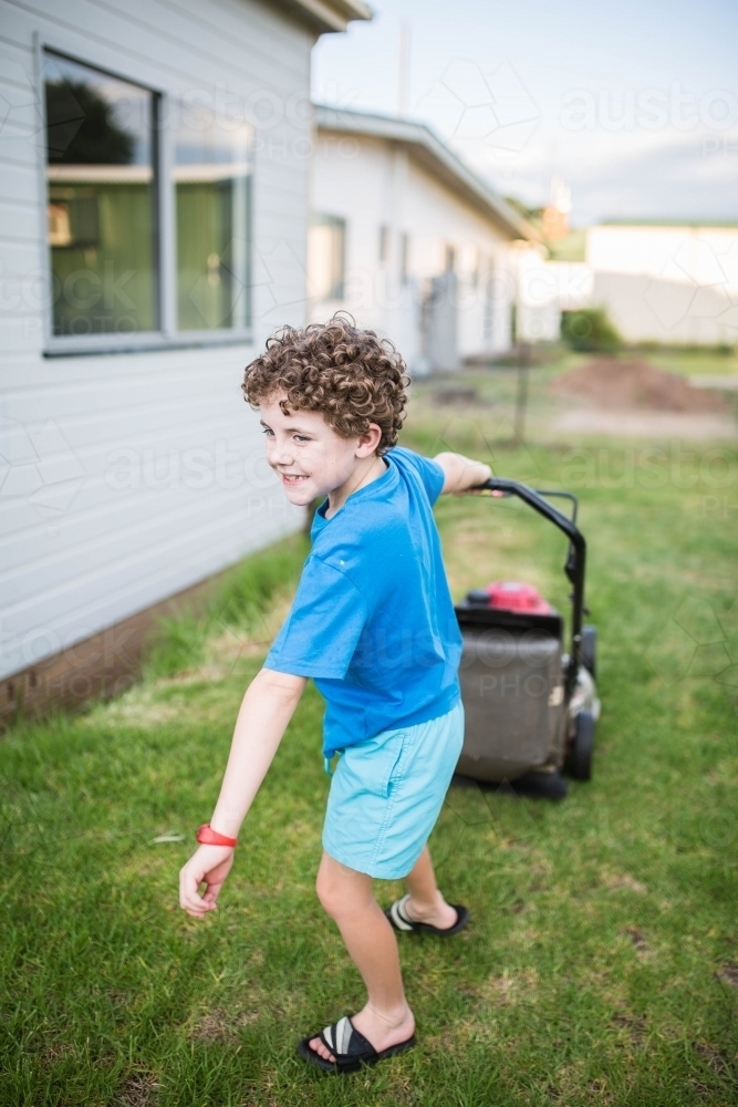 Boy pulling lawn mower mowing lawn smiling - Australian Stock Image