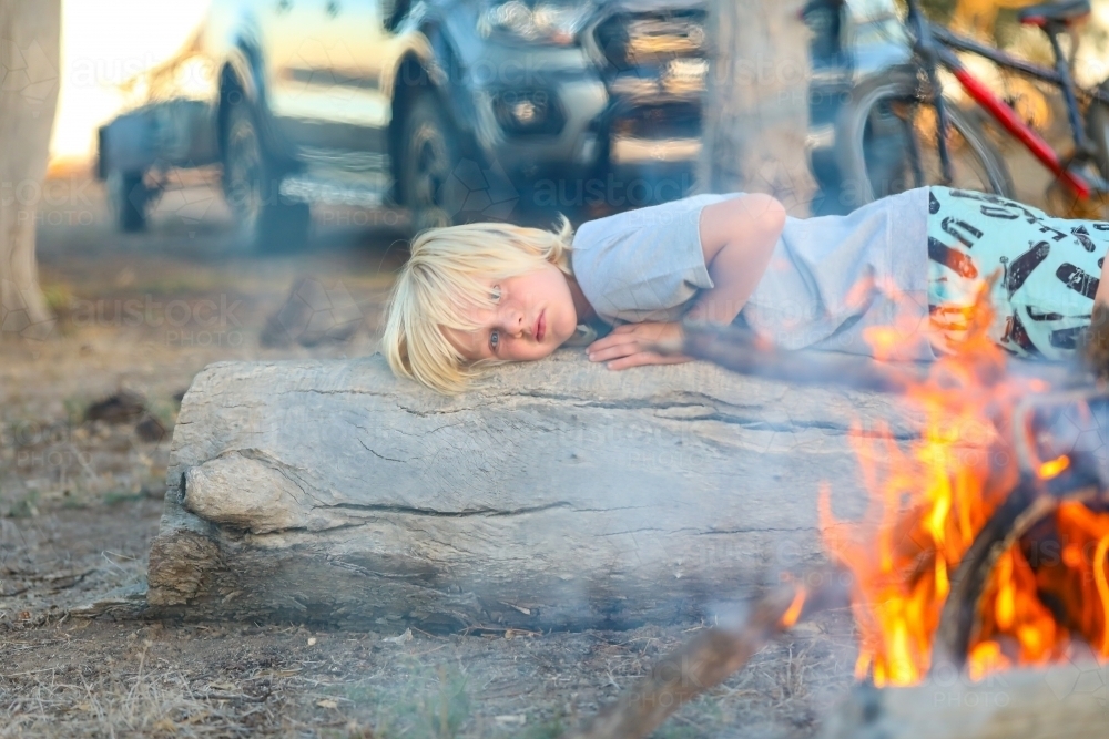 Boy relaxing on log near camp fire - Australian Stock Image