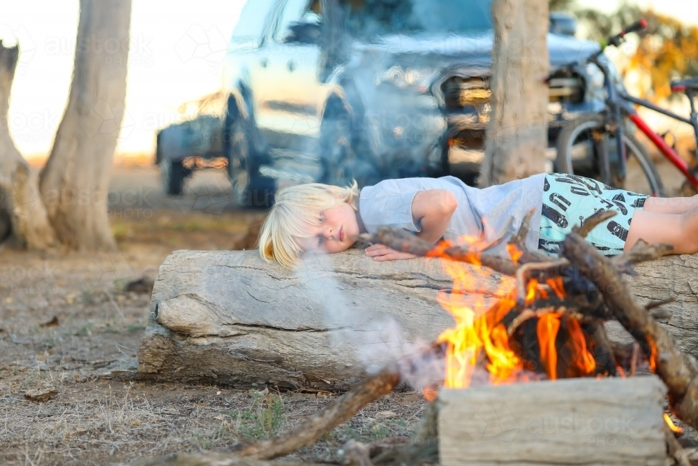 Boy relaxing on log near camp fire - Australian Stock Image