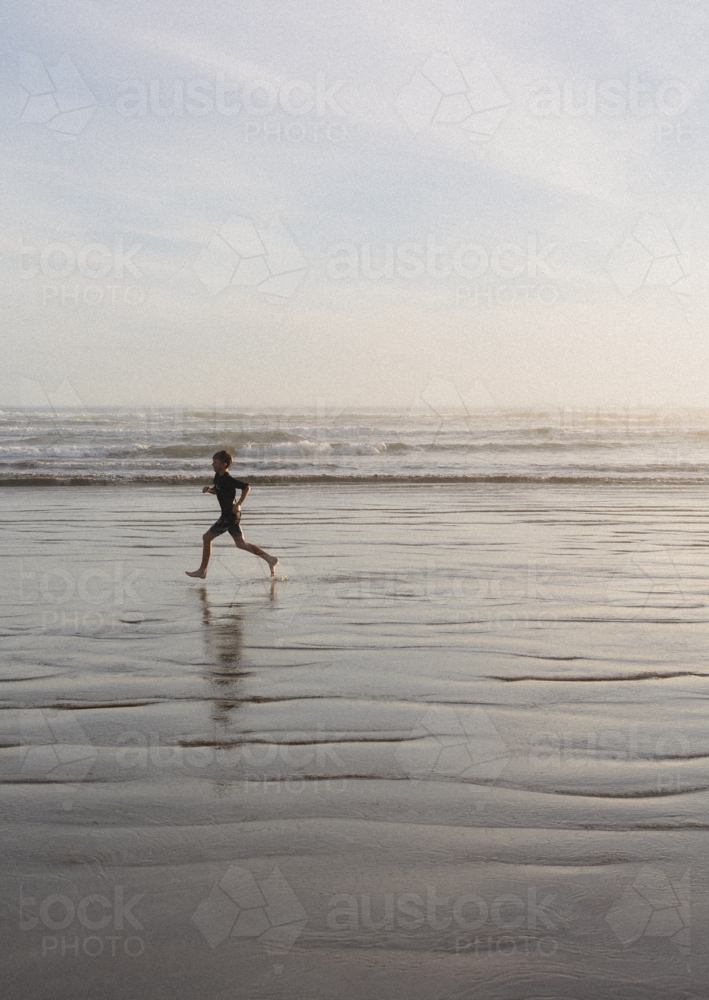 Boy running along remote beach at sunset - Australian Stock Image