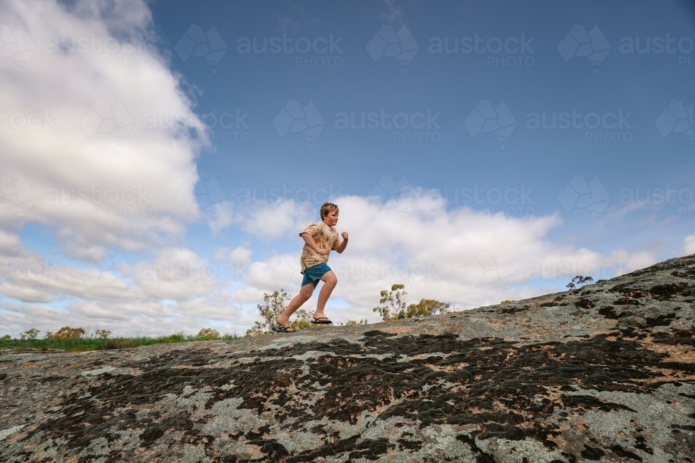 Boy running and jumping on rocks under cloudy blue sky in nature - Australian Stock Image