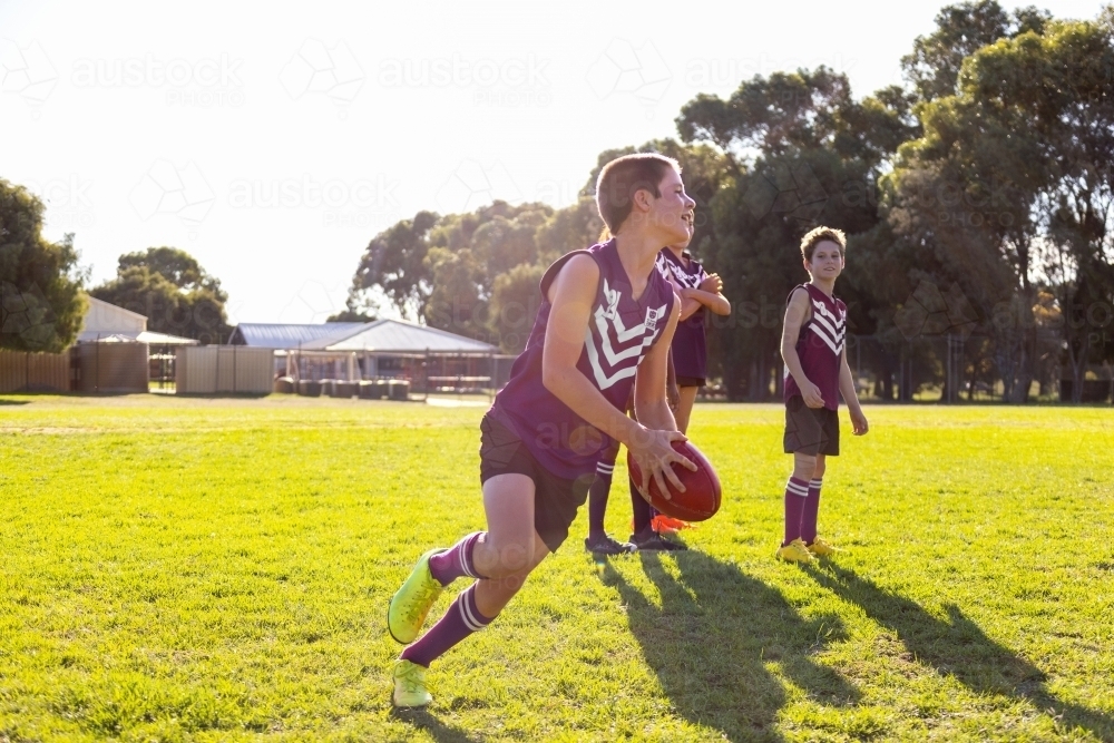 boy running on grass with australian rules football as teammates watch - Australian Stock Image