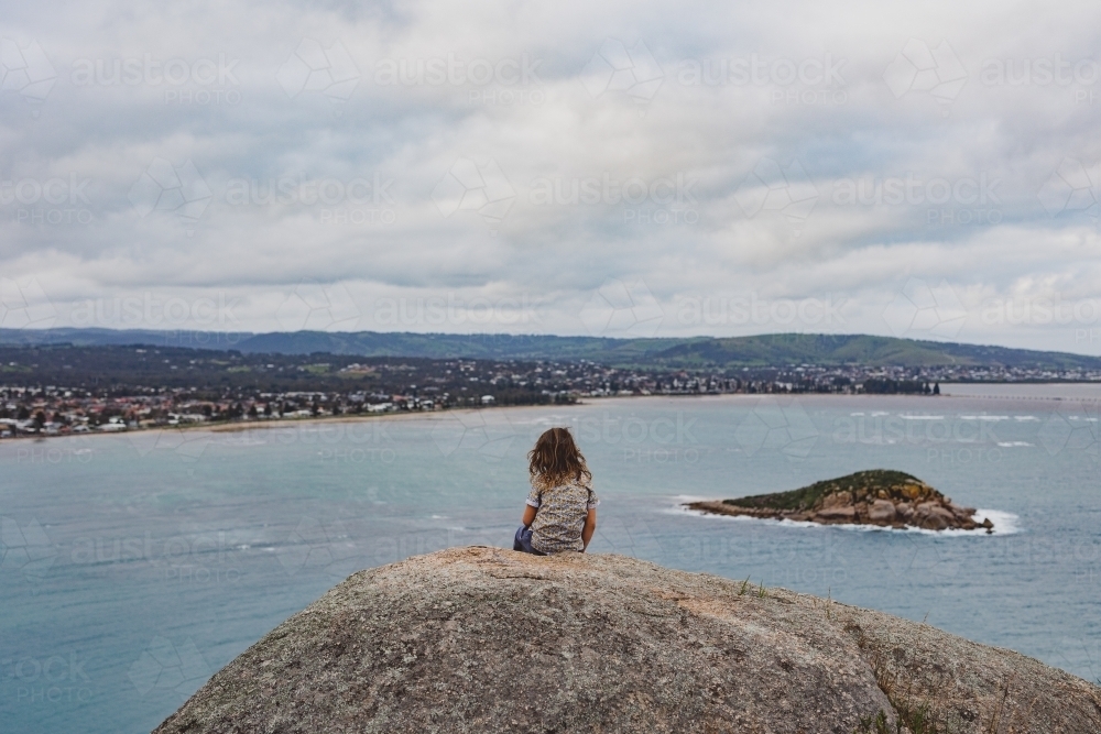 Boy sitting on a rock looking at coastline - Australian Stock Image