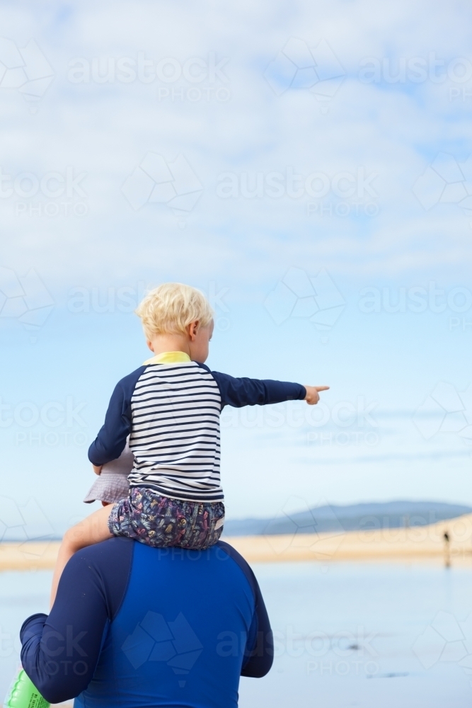 Boy sitting on fathers shoulders at the beach pointing away - Australian Stock Image