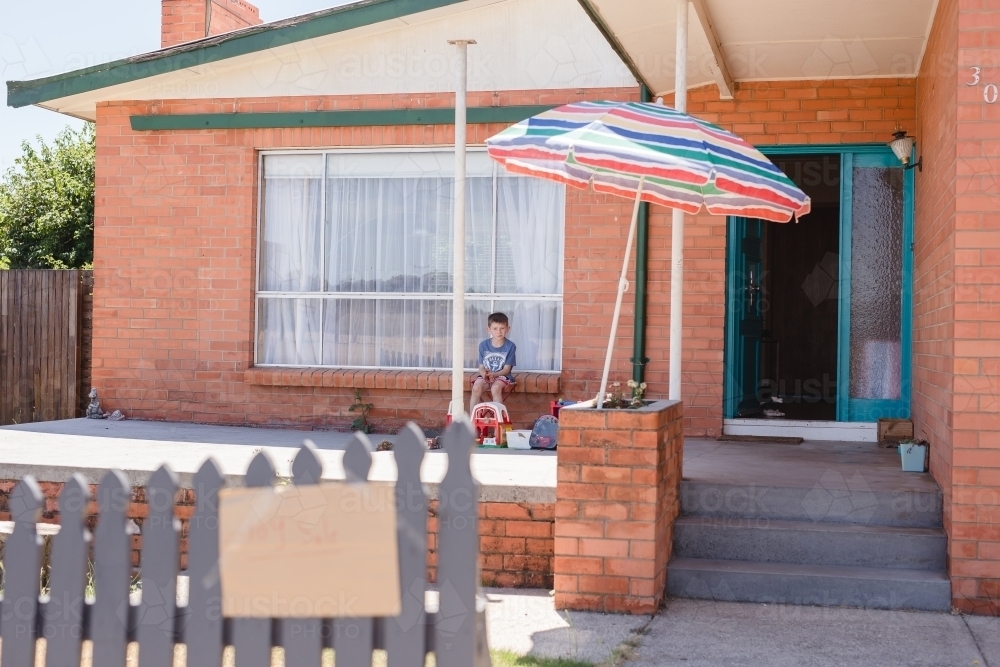 Boy sitting on front porch of home with Toy Sale sign on fence - Australian Stock Image