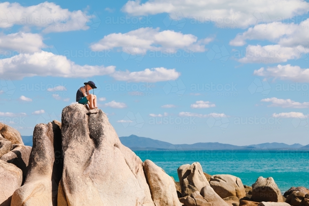 Boy sitting on high rock by the beach in the Whitsundays - Australian Stock Image