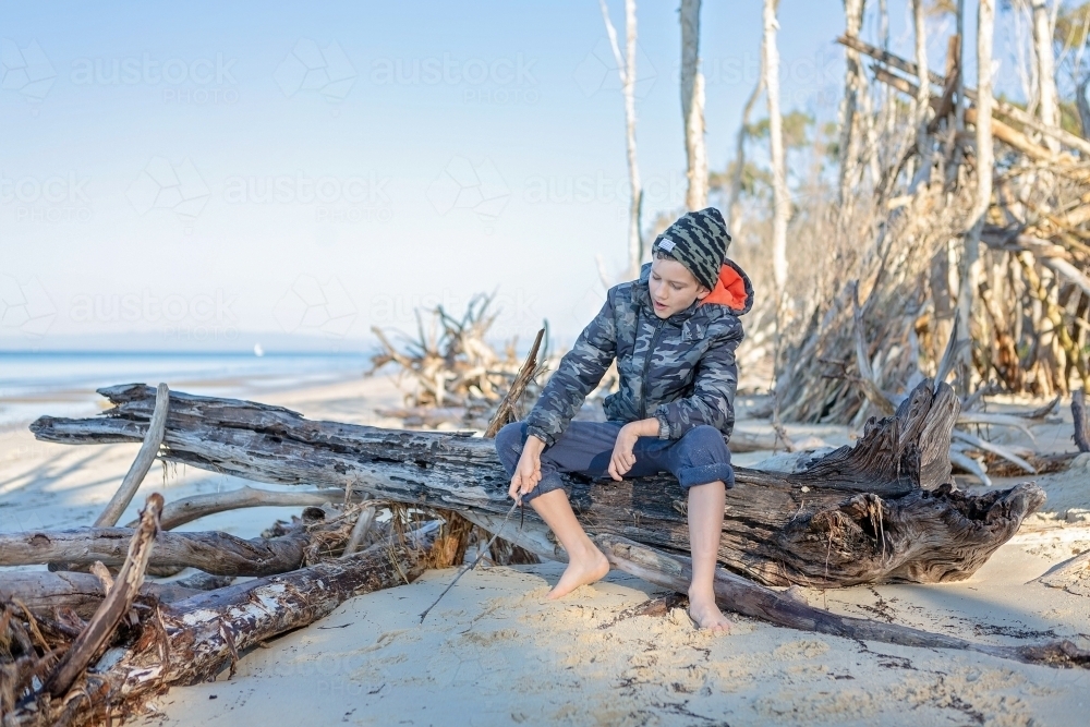 Boy sitting on log on remote beach in Queensland - Australian Stock Image