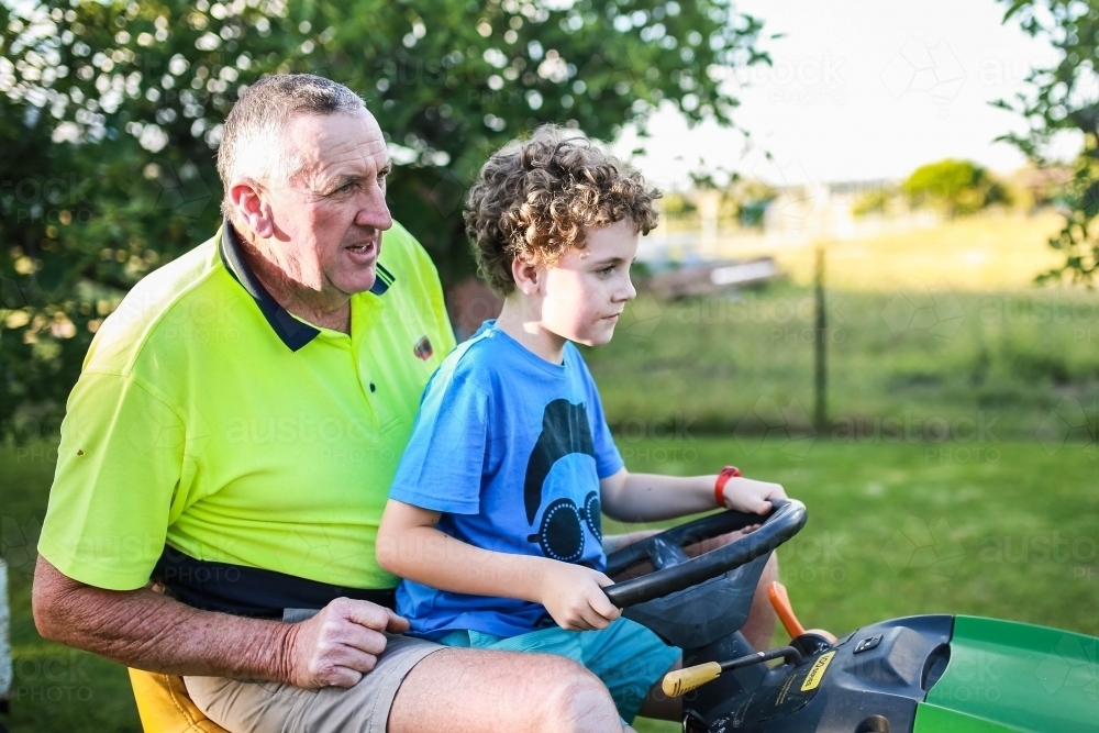 Boy sitting on ride on lawn mower with grandfather - Australian Stock Image