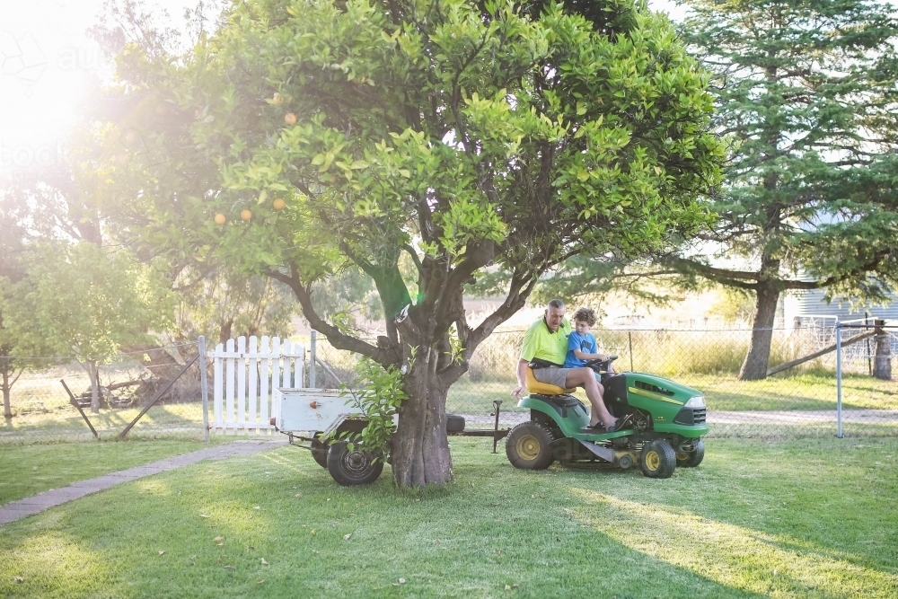 Boy sitting on ride on lawn mower with grandfather towing white trailer - Australian Stock Image