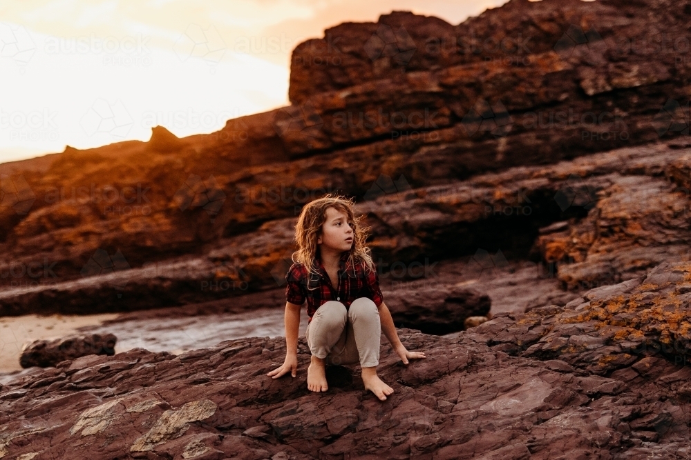 Boy sitting on rocks at the ocean at sunset - Australian Stock Image