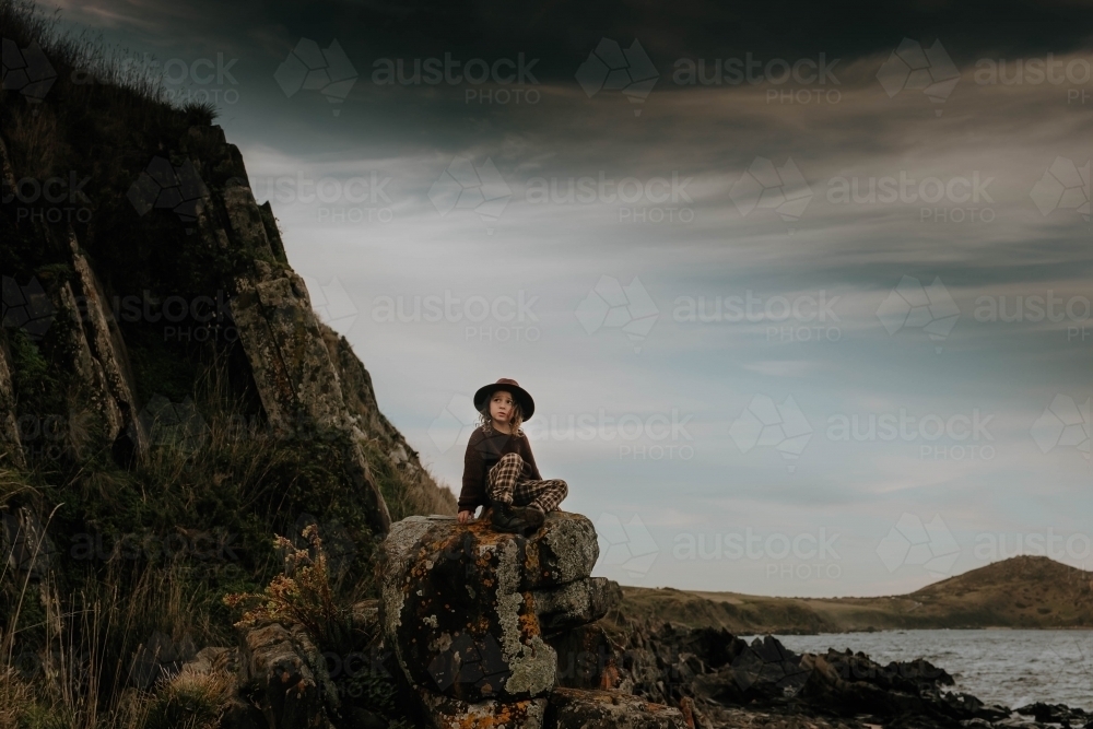 Boy sitting on rocks next to cliffs and ocean a very dark moody sky - Australian Stock Image