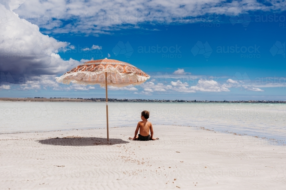 Boy sitting under an umbrella on the sand next to ocean - Australian Stock Image